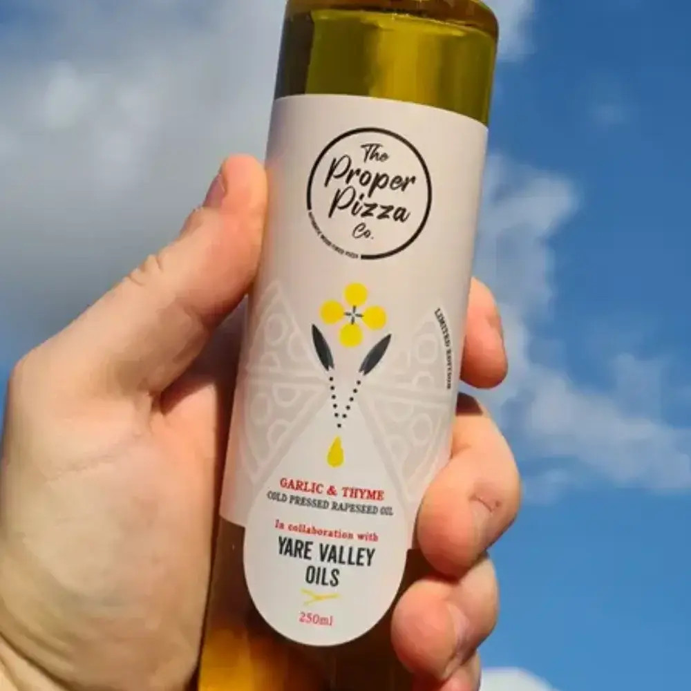 Hand holding a bottle of The Proper Pizza Co garlic and thyme cold pressed rapeseed oil in collaboration with Yare Valley Oils against a blue sky.
