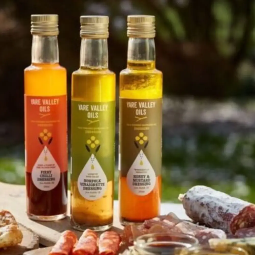 Three Yare Valley dressing bottles displayed outdoors with a selection of food on a rustic table.